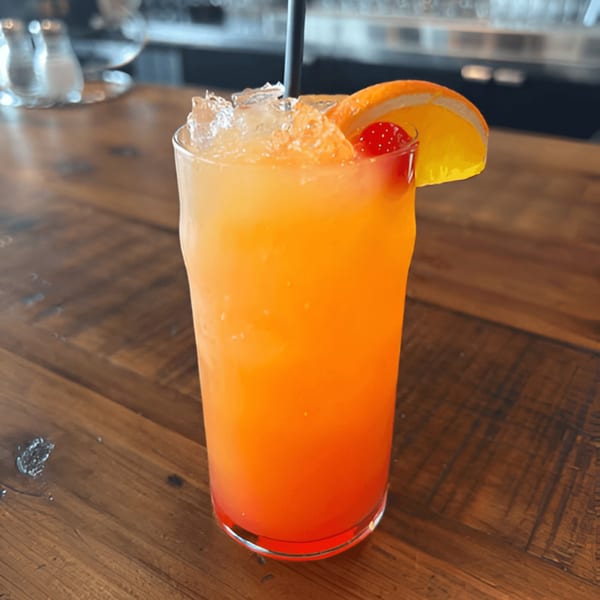 Vibrant orange and red cocktail with ice, an orange slice, and a cherry on a wooden table.