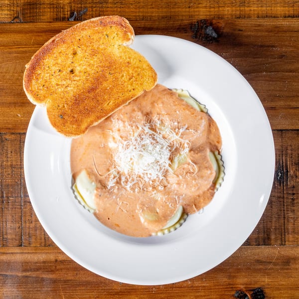 Ravioli with creamy sauce and shredded cheese, served with a slice of garlic bread on a white plate.