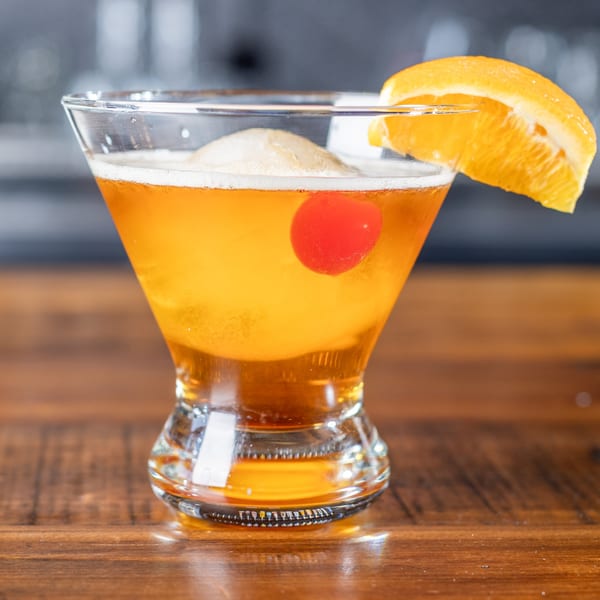 A Wicked Peel Pizza cocktail with an orange slice and cherry on a wooden table.