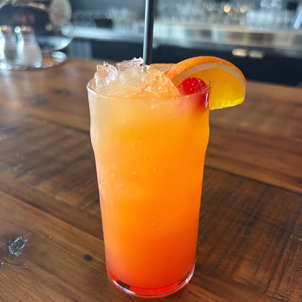 A vibrant orange and red cocktail with ice, a black straw, orange slice, and cherry on a wooden table.