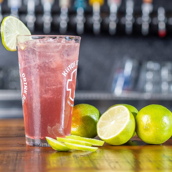 A pink drink with ice and a lime wedge, surrounded by whole and sliced limes on a wooden surface.